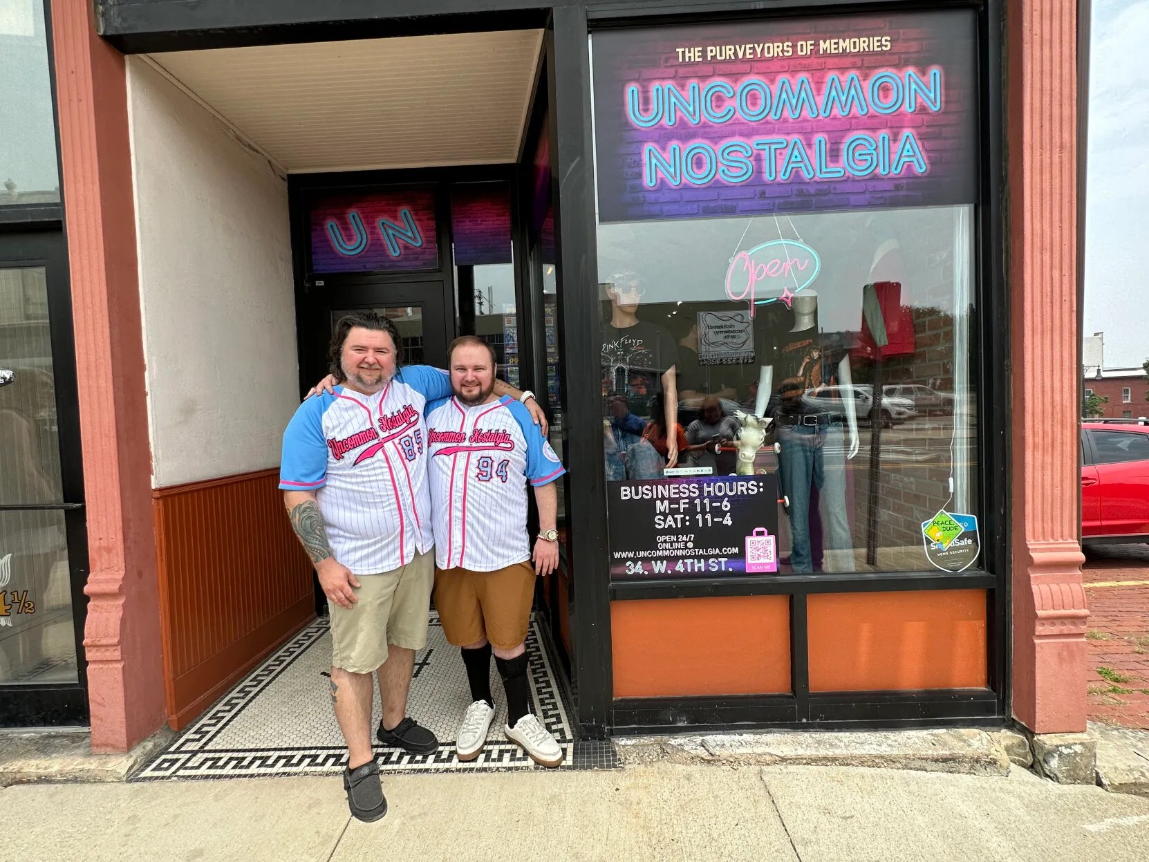 Two men stand in baseball jerseys in front of their store with a pink and blue sign.
