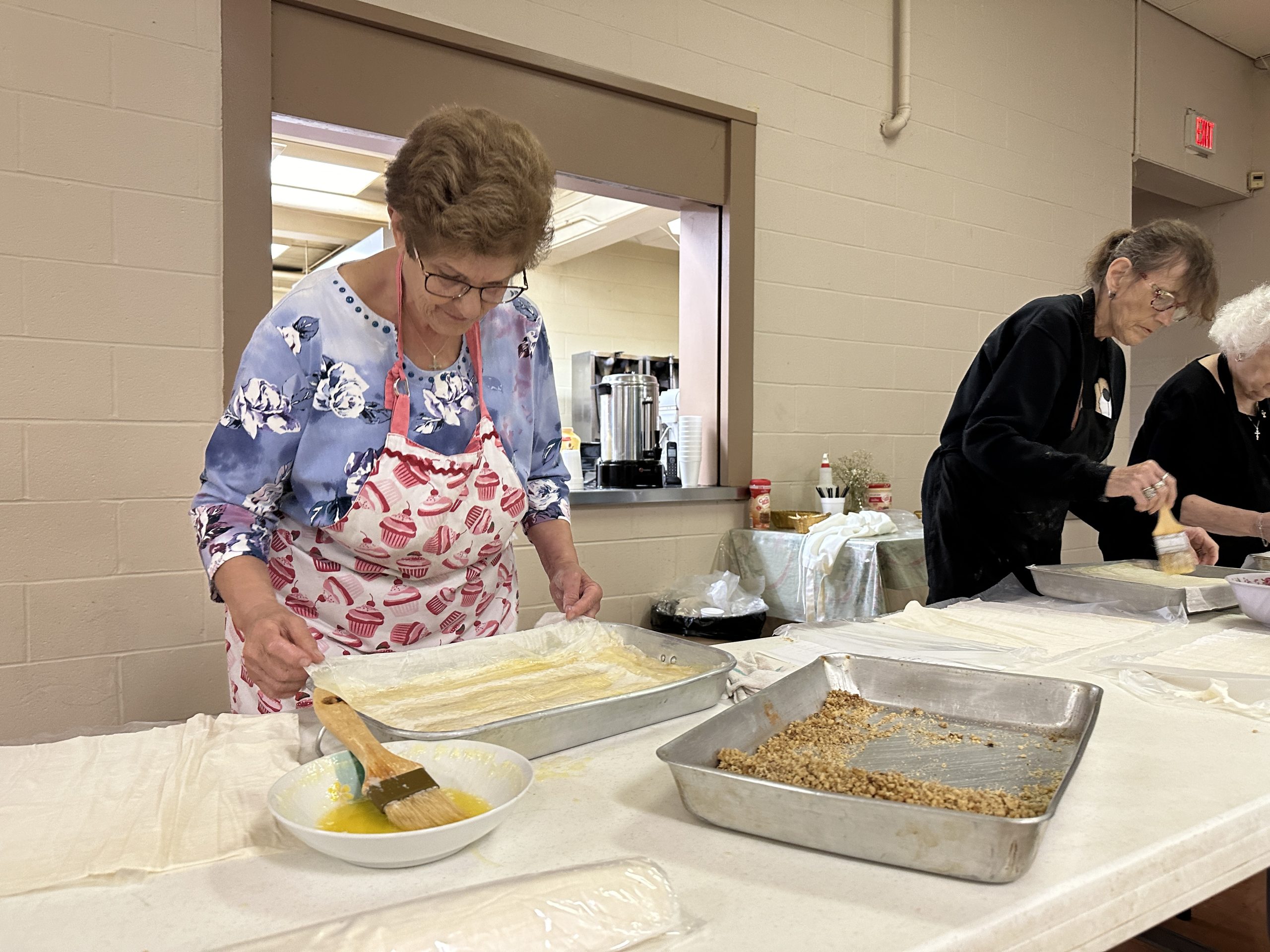 Older woman coats a brush in a bowl of sugar syrup in order to continue coating the philo dough in front of her. She is preparing baklava in the kitchen of the Sts. Constantine and Helen Greek Orthodox Church for the 2024 Greek Festival.