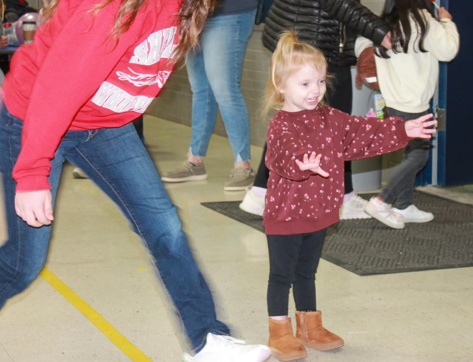 A young girl wearing a red sweater with pink flowers on it smiles with her arms open at a Richland Outreach Center event.