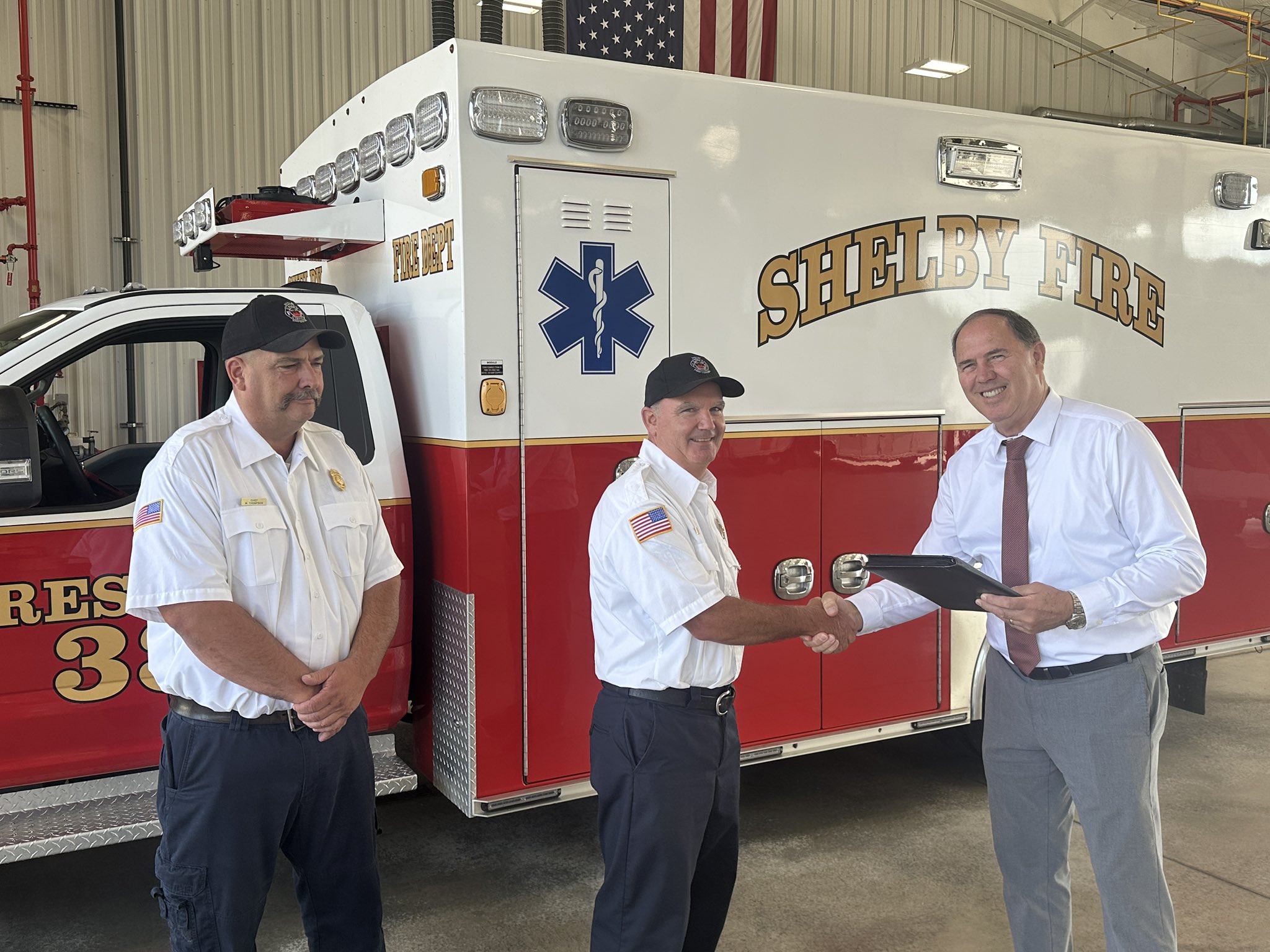 Two firemen standing with city mayor in fire station by ambulance