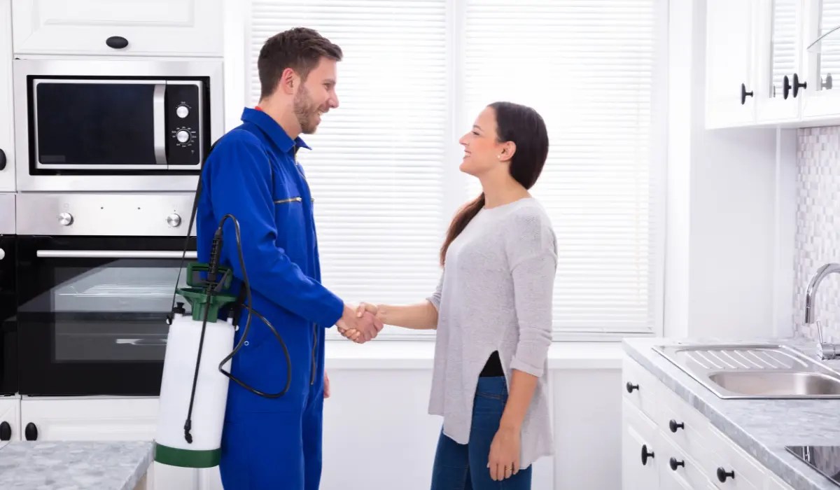 Pest control technician in blue uniform shaking hands with a smiling homeowner in a bright white kitchen