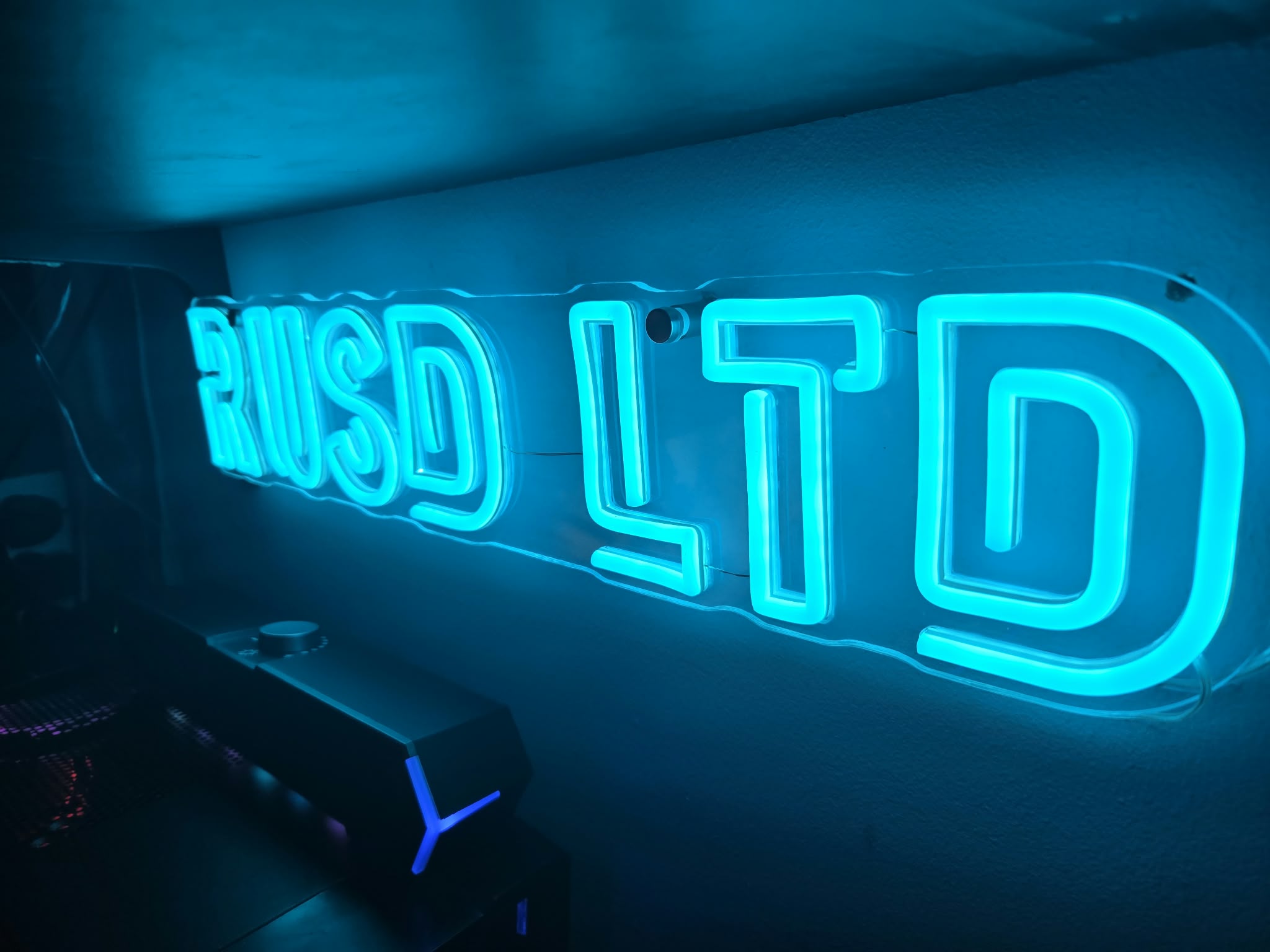 Blue neon sign with glowing letters mounted on a dark wall in a dim room (partially visible text).