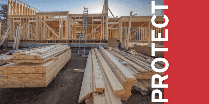 Under-construction wooden framing of a building with stacked lumber on a dirt site (blue sky).