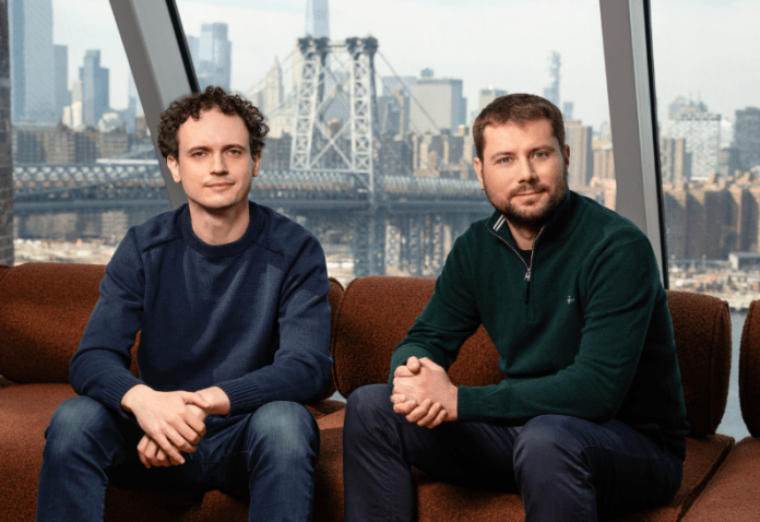 Two men-reflection ai partners, one in a blue sweater and the other in a dark green sweater, sit on a couch in a modern office. A large window behind them overlooks the New York City skyline, including the Manhattan Bridge.