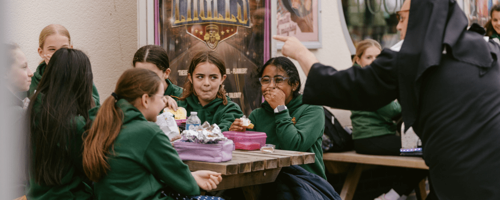 A group seven children sitting at bench eating their packed lunches, laughing as Musical Ruth speaks to them
