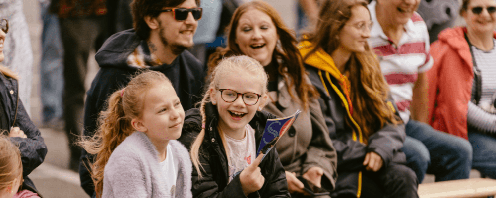 A close up of the audience at Mablethorpe. Two children, one laughing, holding the SO Festival programme, the second looking pensive, with a group of adults laugh at the performance