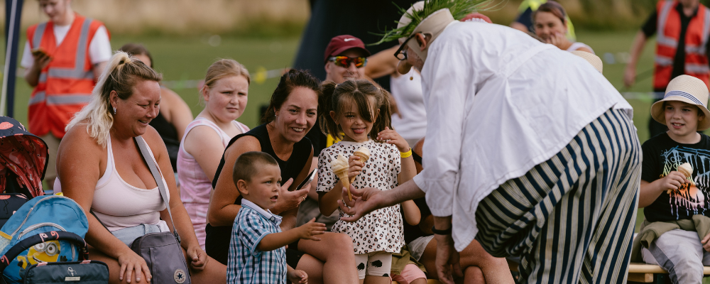 A group of children and adults smiling and watching as a clown in stripy trousers and green hair serves ice cream cones to them