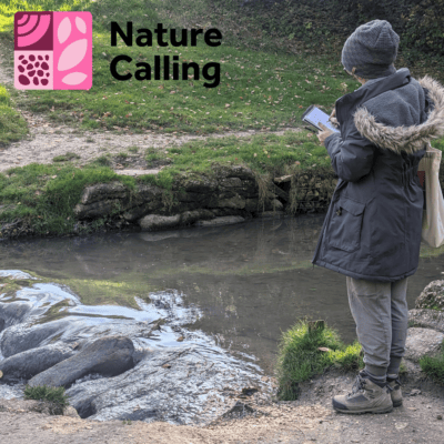 A person stood next to a stream sketching, wearing a coat, woolly hat and walking boots.