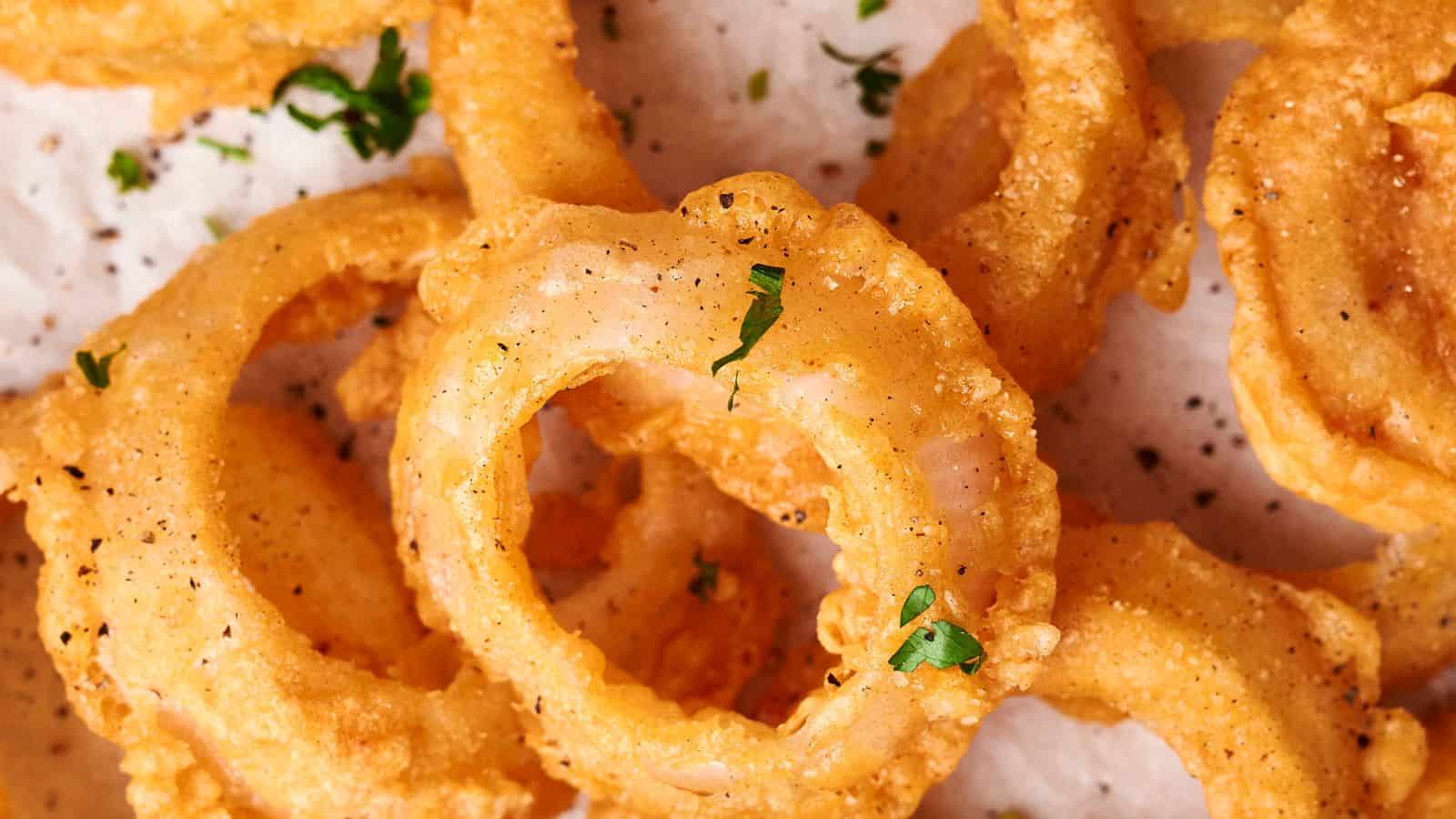 A close-up of crispy, golden-brown onion rings sprinkled with chopped parsley and black pepper on a white parchment-lined surface captures perfection in every bite.