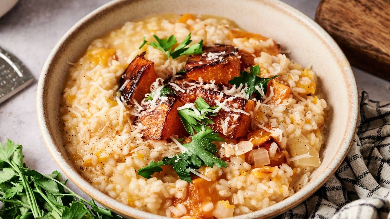A bowl of risotto topped with roasted pumpkin pieces, grated cheese, and fresh parsley, with a checkered napkin and herbs nearby.