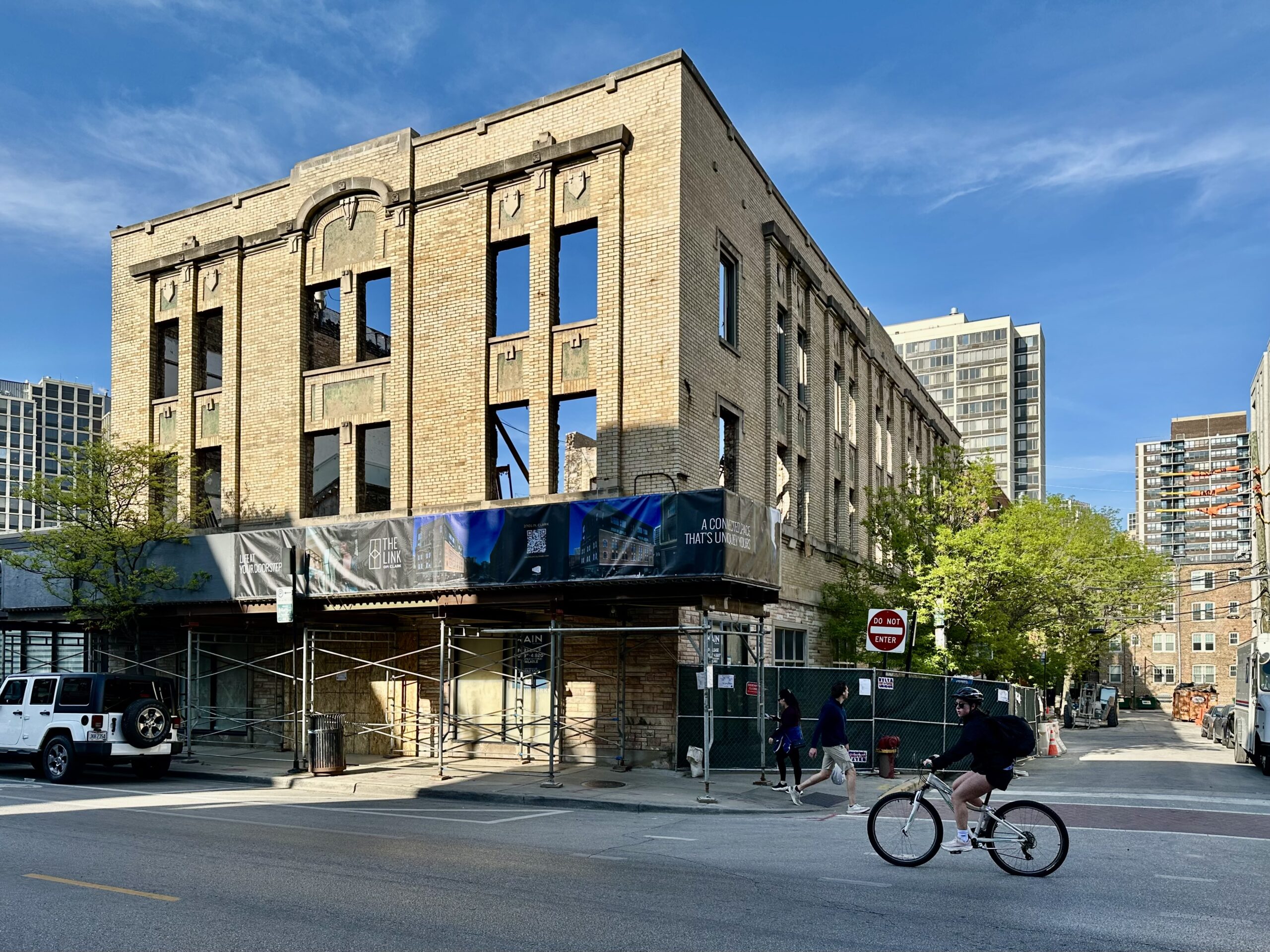 A three-story tan brick building with decorative stone trim on a street corner, gutted for renovation. Its upper windows are empty openings with blue sky visible through them. Scaffolding and a construction banner reading "The Link on Clark" wrap the ground floor, and a cyclist rides past on Clark Street.