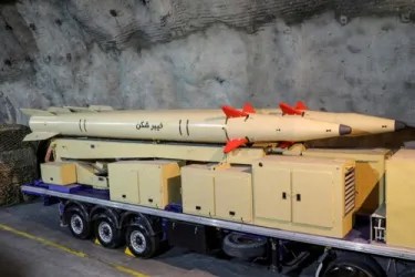 Military missile transport trailer carrying three beige missiles with red fins on a multi-axle bed inside a hangar.
