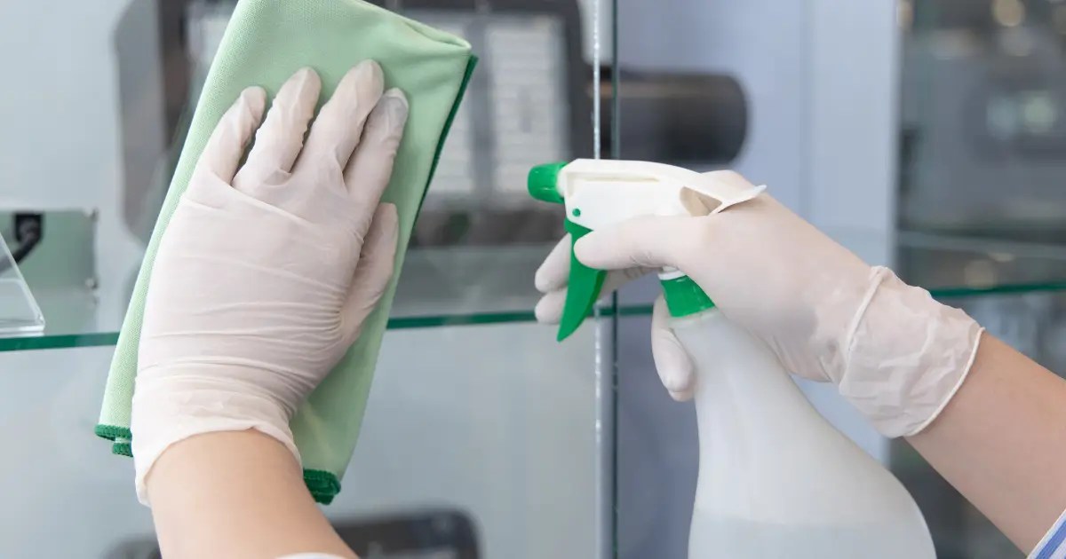 employee with gloves spraying cleaning product on glass with towel in hand