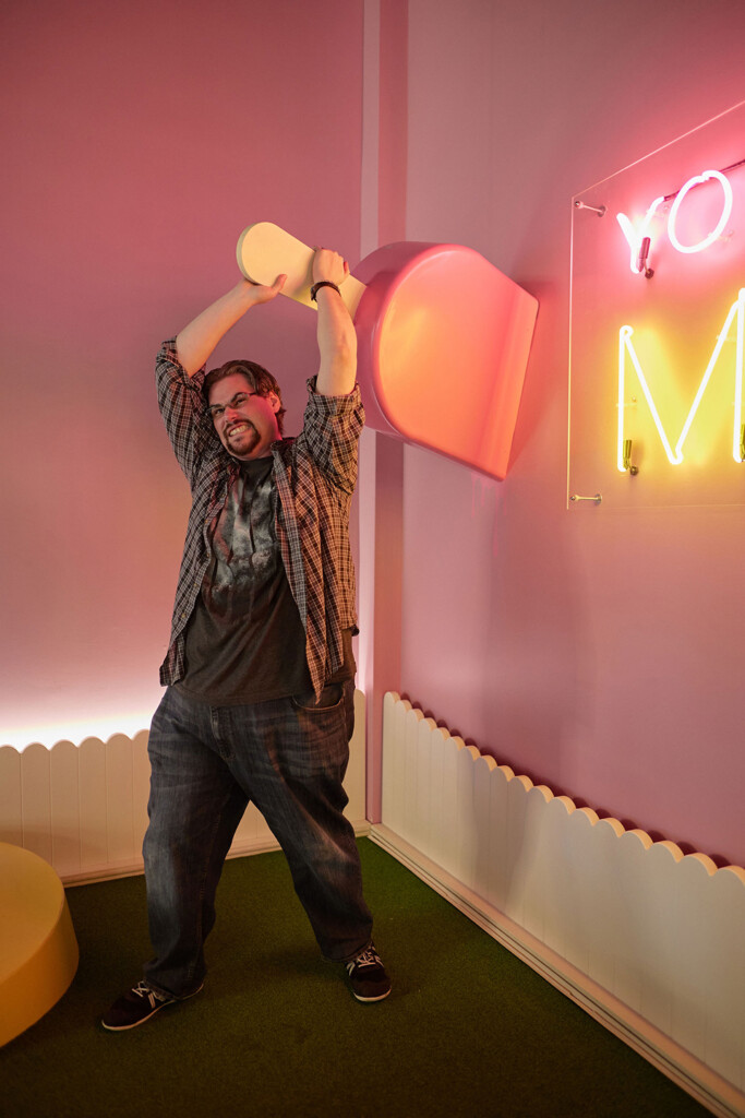 A person is playfully posing with a large, colorful scoop, raised above their head as if about to throw it. They are smiling and wearing a plaid shirt over a graphic t-shirt, paired with loose jeans and sneakers. The background features a pink wall with a neon sign that reads "YOU" in bright colors. The floor is covered in green grass-like material, and there is a circular, yellow platform nearby.