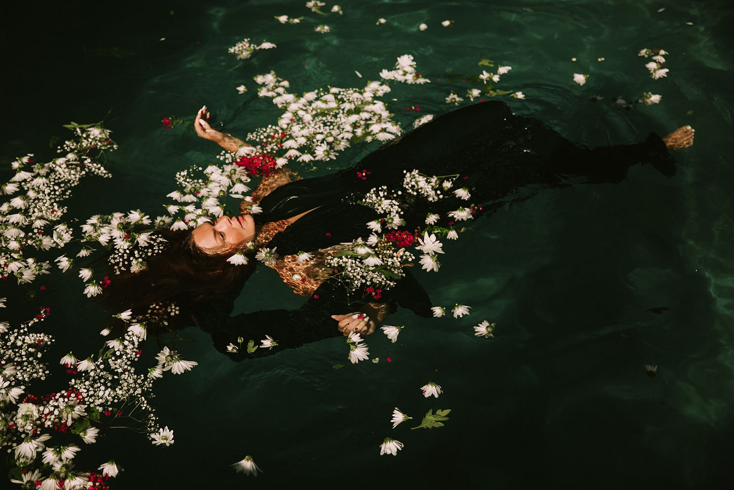 Woman in a black dress floating in water, surrounded by red and white flowers