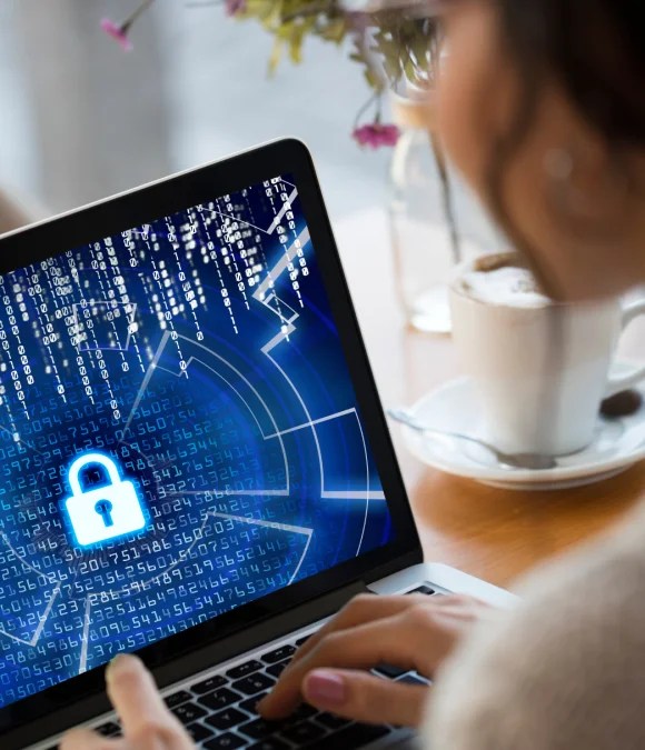 A person seen from over their shoulder is typing on a laptop. The screen displays a cybersecurity graphic with a large, glowing blue padlock icon over streams of digital code.