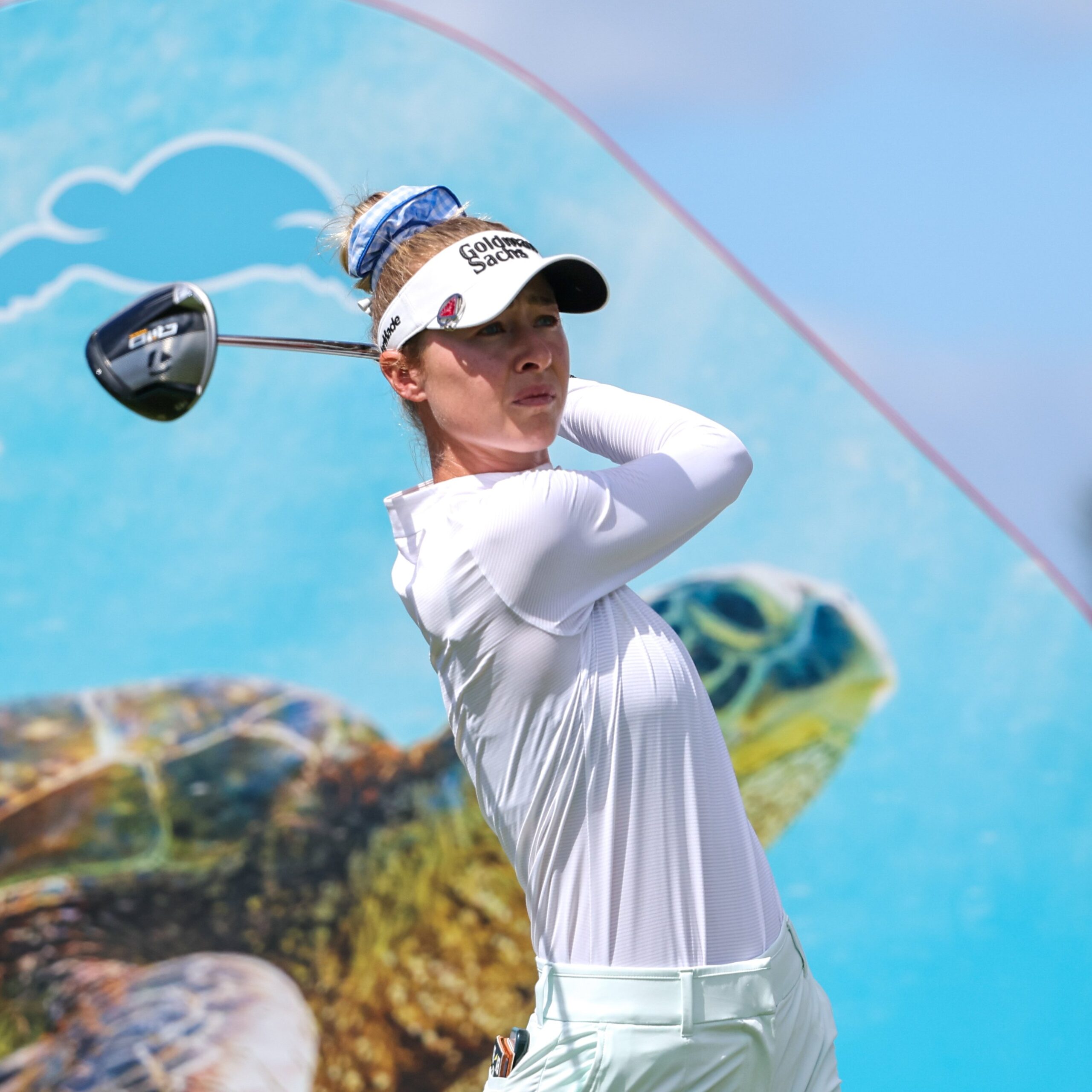 Nelly Korda follows through on a golf swing. The head of the club is to the left of her head, in a follow-through position. Tournament signage for the 2025 LOTTE Championship is behind her: an image of a sea turtle on an aqua blue background.