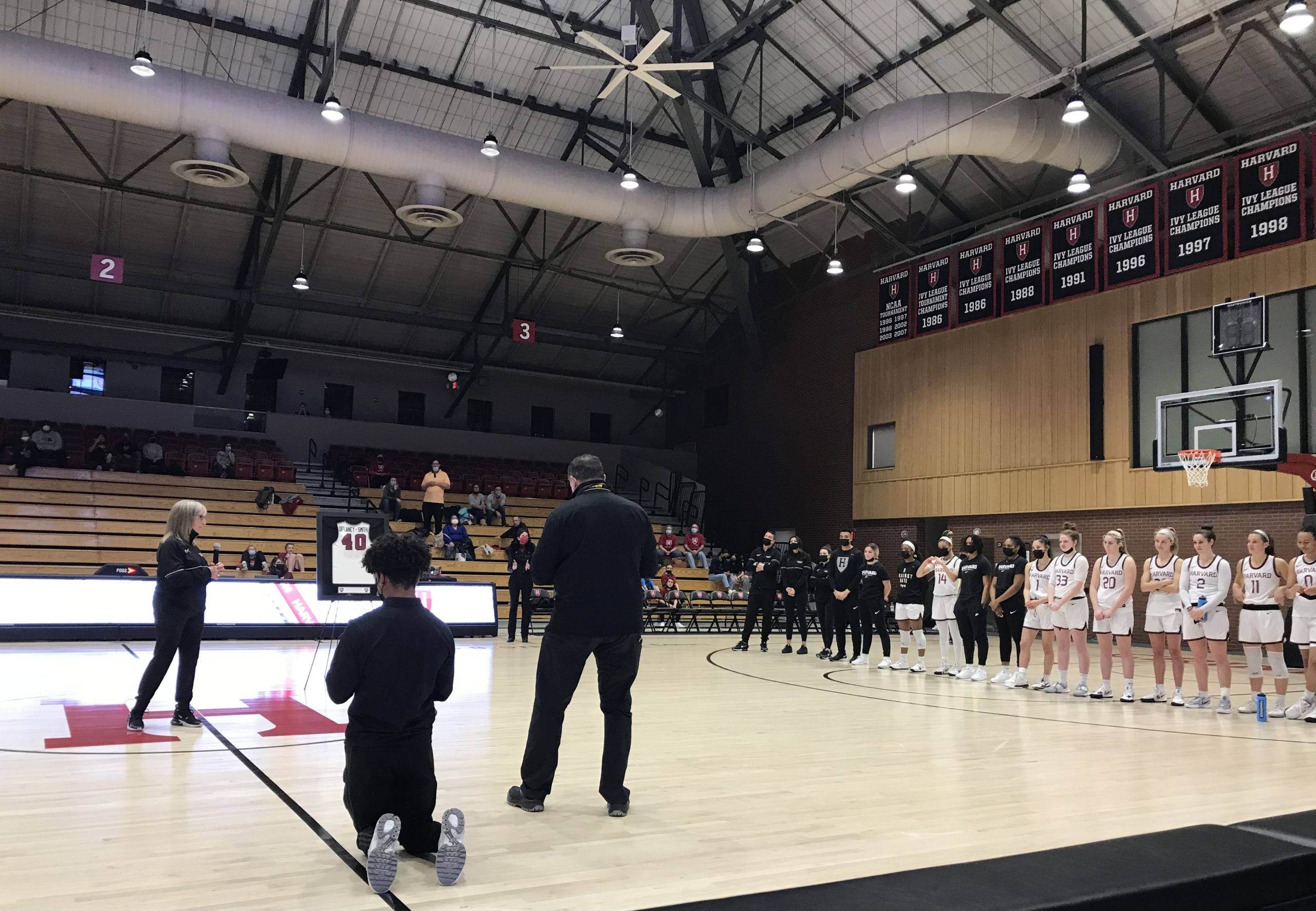Harvard head coach Kathy Delaney-Smith (left) addresses her team during a postgame ceremony on "KDS Day" on Feb. 12, 2022, at Lavietes Pavilion in Cambridge, Massachusetts. Freshman guard Harmoni Turner (14) forms a heart with her hands, and above the players in the rafters are some of Harvard's championship banners under Delaney-Smith. (Photo credit: Jenn Hatfield)