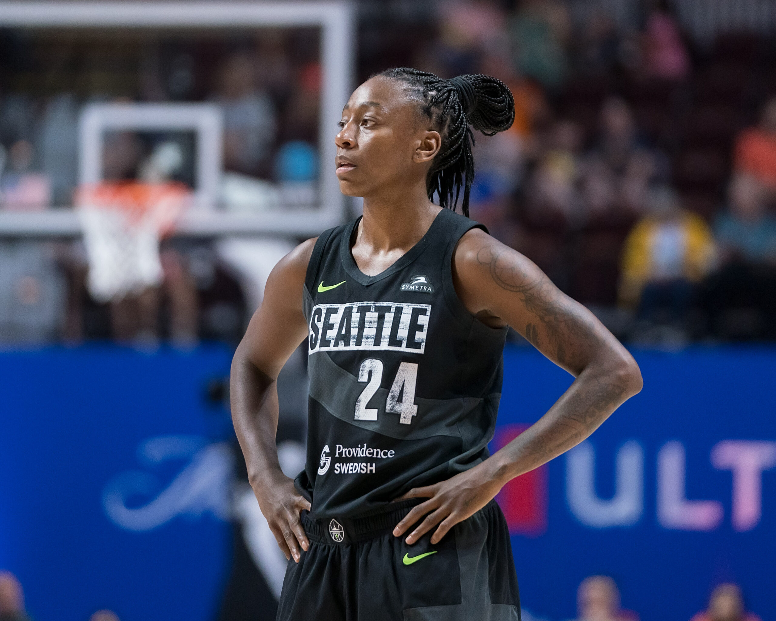 Jewell Loyd stands with a basketball hoop in the distance, wearing Seattle's black uniform, with hands on hips.