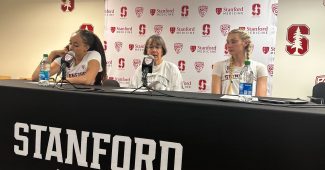 Stanford head coach Tara VanDerveer sits at the podium and speaks at a press conference. Two of her players are on either side of her, and a backdrop of Stanford and Pac-12 logos is displayed behind them.