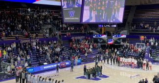 The University of Washington Women's Basketball team stands in a line along the left side free throw line of the Alaska Airlines Arena facing the Washington State Women's Basketball aligned on the right side free throw line during the National Anthem being sung by a group of elementary school children on Jan 14, 2024 at Alaska Airlines Arena in Seattle. The singers are visible on a large jumbotron screen above the basketball court.