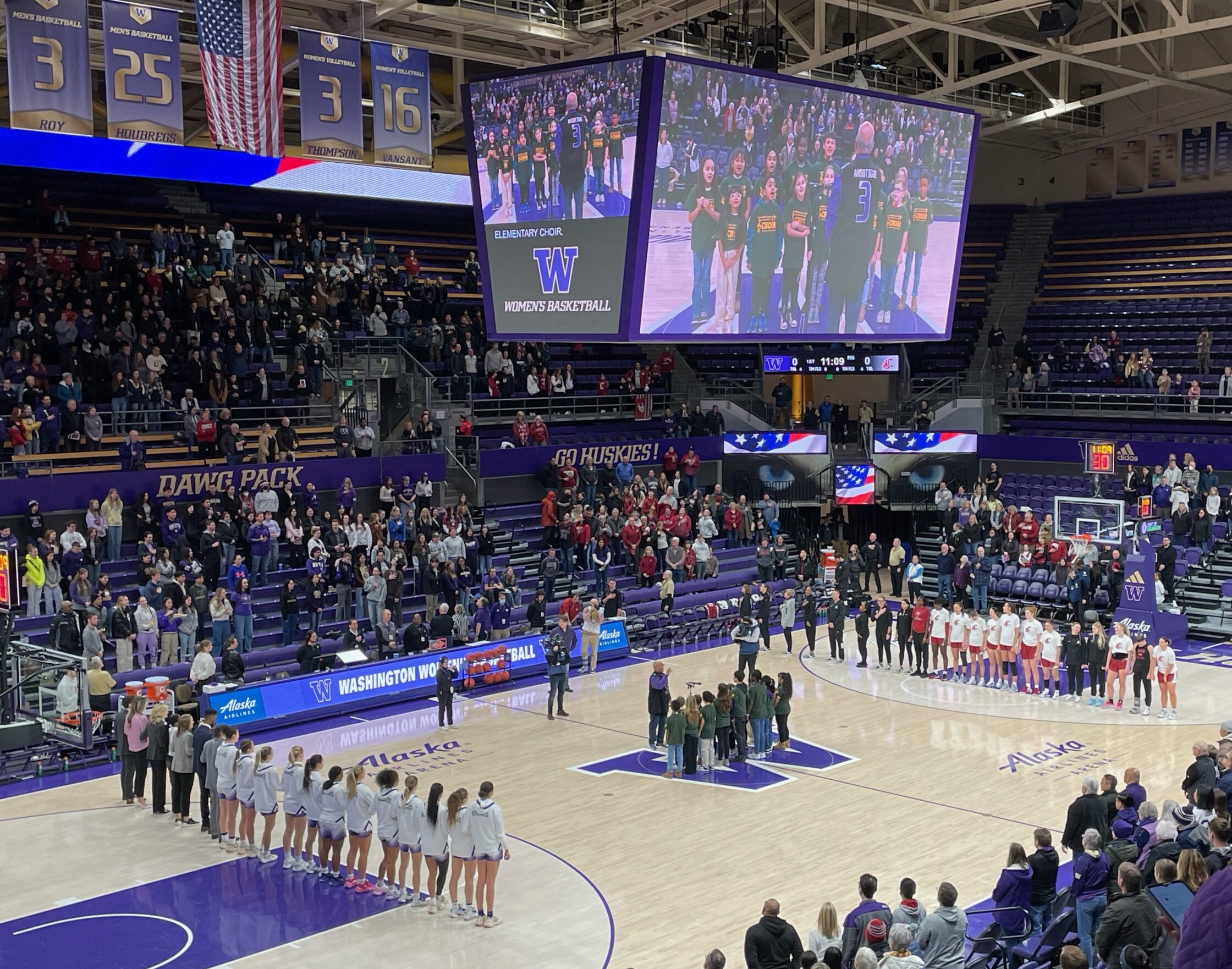 The University of Washington Women's Basketball team stands in a line along the left side free throw line of the Alaska Airlines Arena facing the Washington State Women's Basketball aligned on the right side free throw line during the National Anthem being sung by a group of elementary school children on Jan 14, 2024 at Alaska Airlines Arena in Seattle. The singers are visible on a large jumbotron screen above the basketball court.