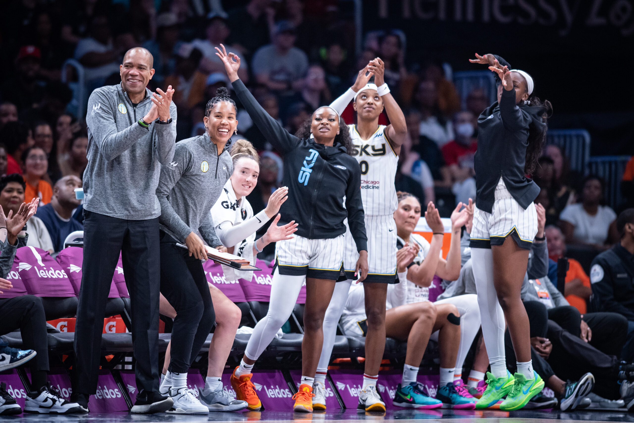 The Chicago Sky bench celebrates a play during a game. Several players stand and clap or make 3-point signs with their hands, and two assistant coaches smile and stand.