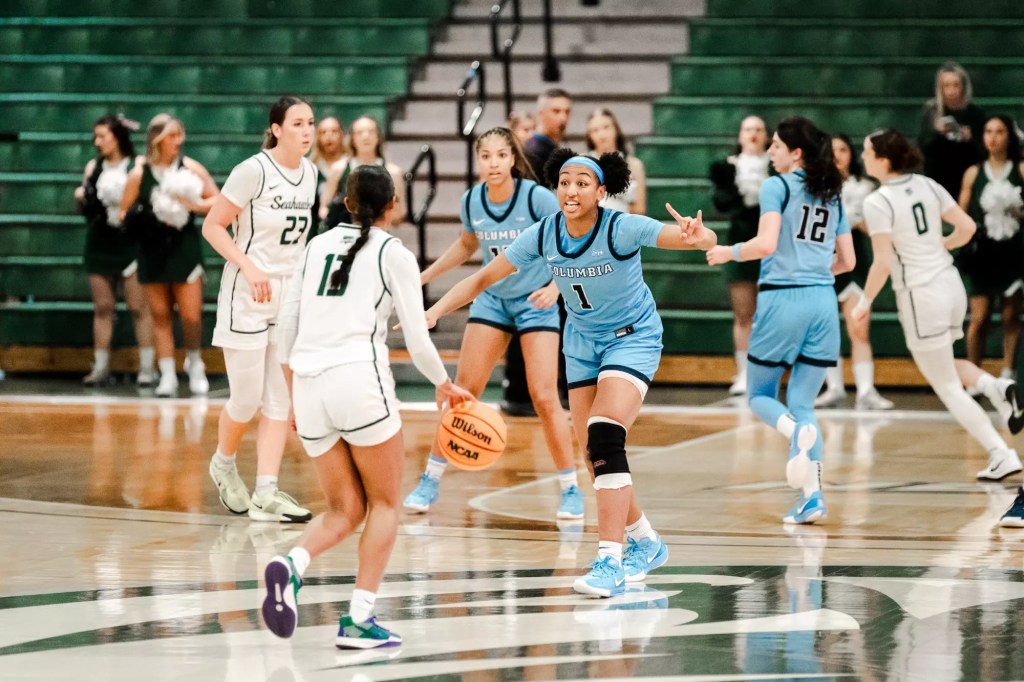 Columbia guard/forward Perri Page stands on the large halfcourt logo, ready to defend the Wagner ball-handler. She holds her left hand up, making a signal that looks like the Texas Longhorns' "Hook 'em Horns" sign.