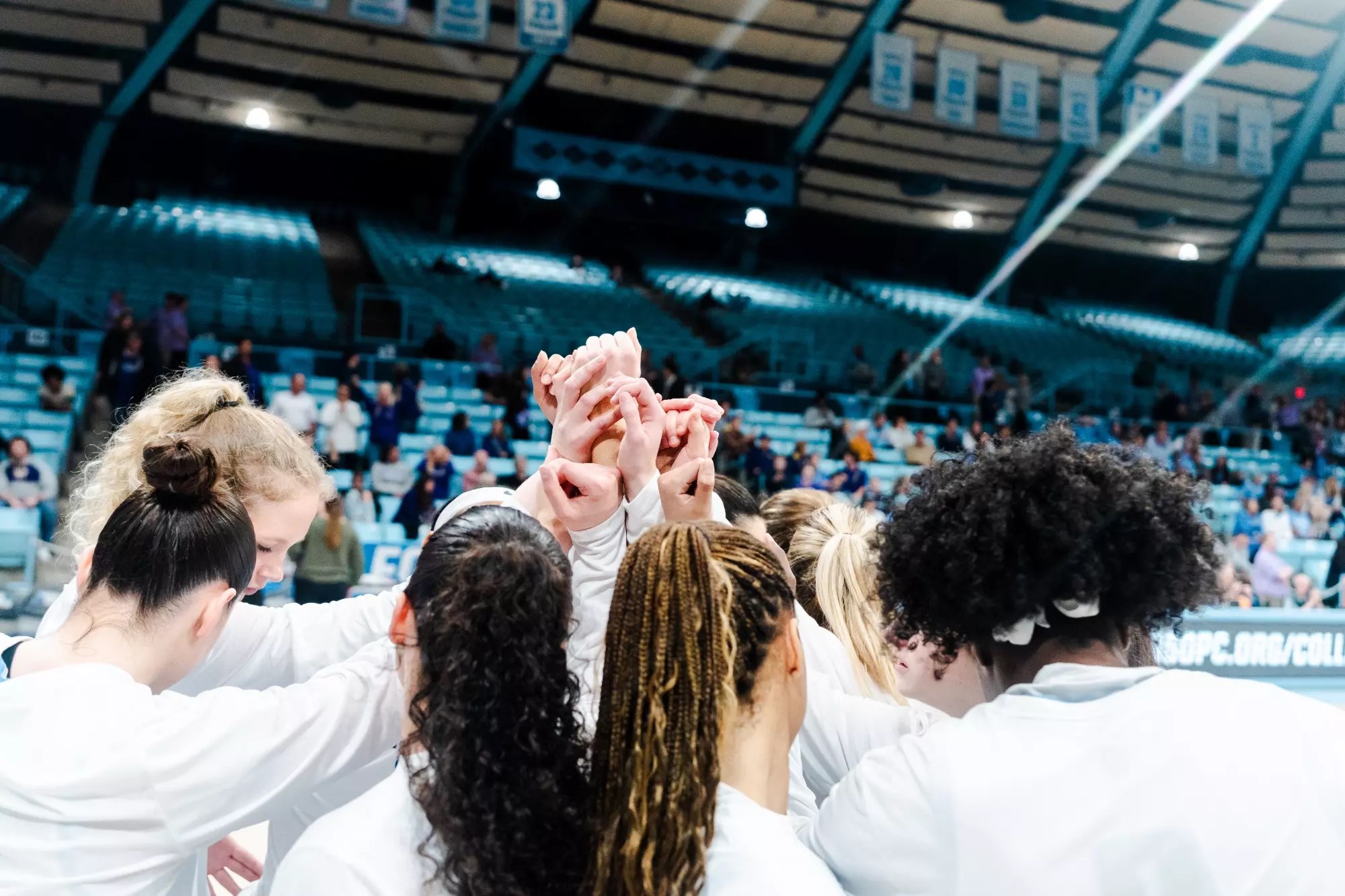 Columbia players are wearing white warmup shirts as they stand in a huddle. They each extend an arm up and toward the center of the circle.