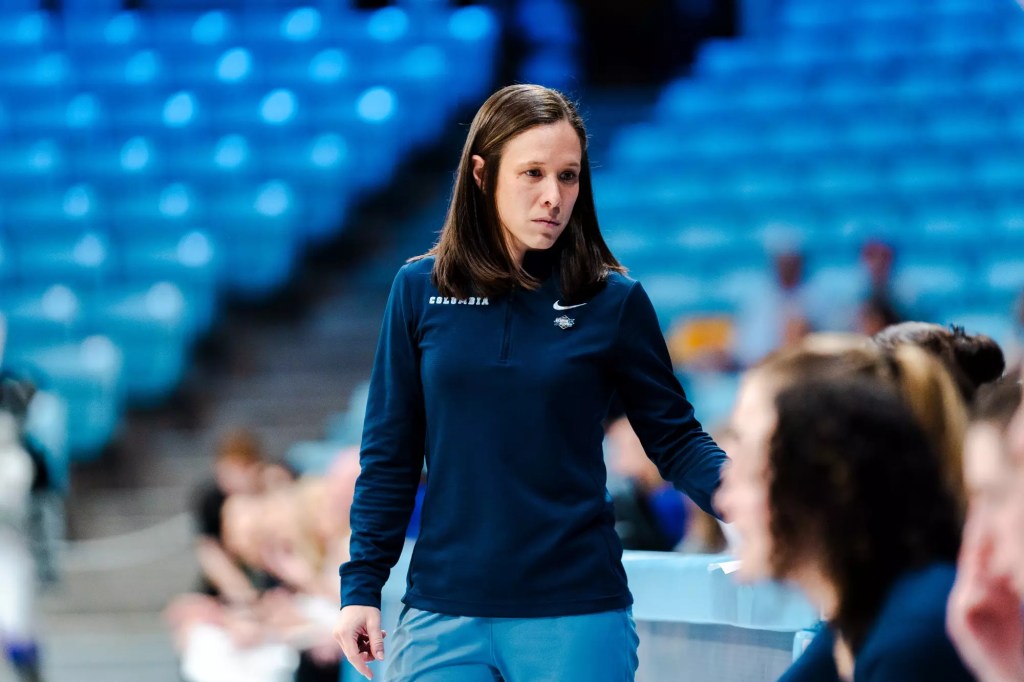 Columbia head coach Megan Griffith wears a dark blue quarter-zip and looks at the players on her bench during a game. She isn't smiling, but she isn't obviously angry, either.