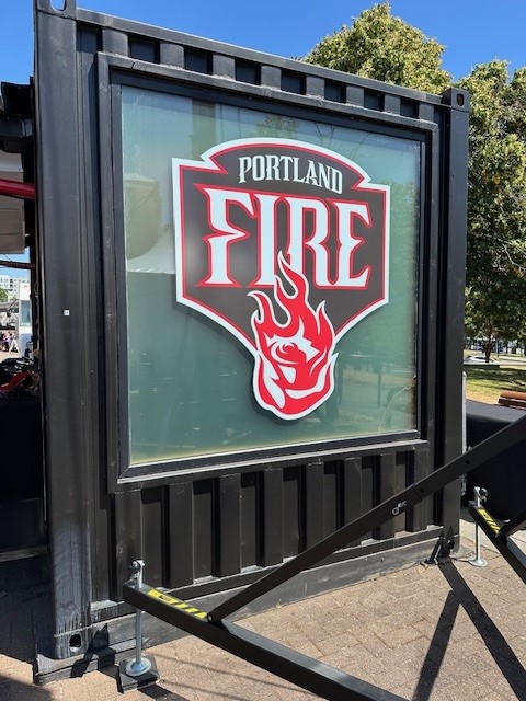 Photo of a large outdoor sign with the Portland Fire logo on it. Logo is all-caps white text with red outline. "Portland" on top in smaller text, with "Fire" below in larger text. flaming red and white rose below. blue background in an upside-down pentagon shape.