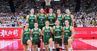 Two rows of Australian basketball players take a team photo before a game. They are on the free throw line with the rim and crowd in the background.