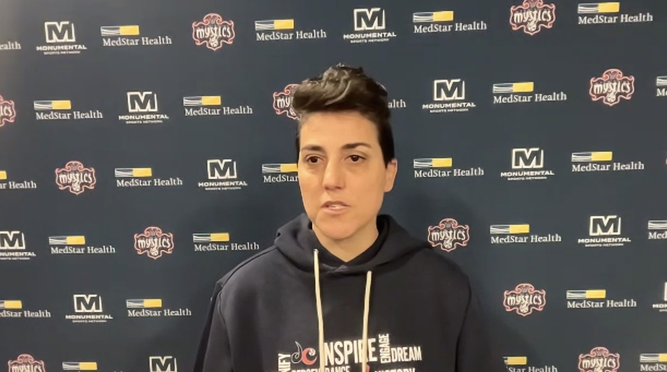 Washington Mystics general manager Jamila Wideman is shown doing a Zoom call with reporters. She is wearing a navy Mystics sweatshirt, and there is a navy backdrop with the Mystics, Monumental Sports Network and MedStar Health logos behind her.
