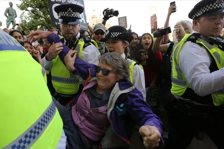 London Protest Crackdown: Police officers detain an older protester wearing sunglasses during a tense demonstration near Parliament, as crowds shout and hold signs in the background.