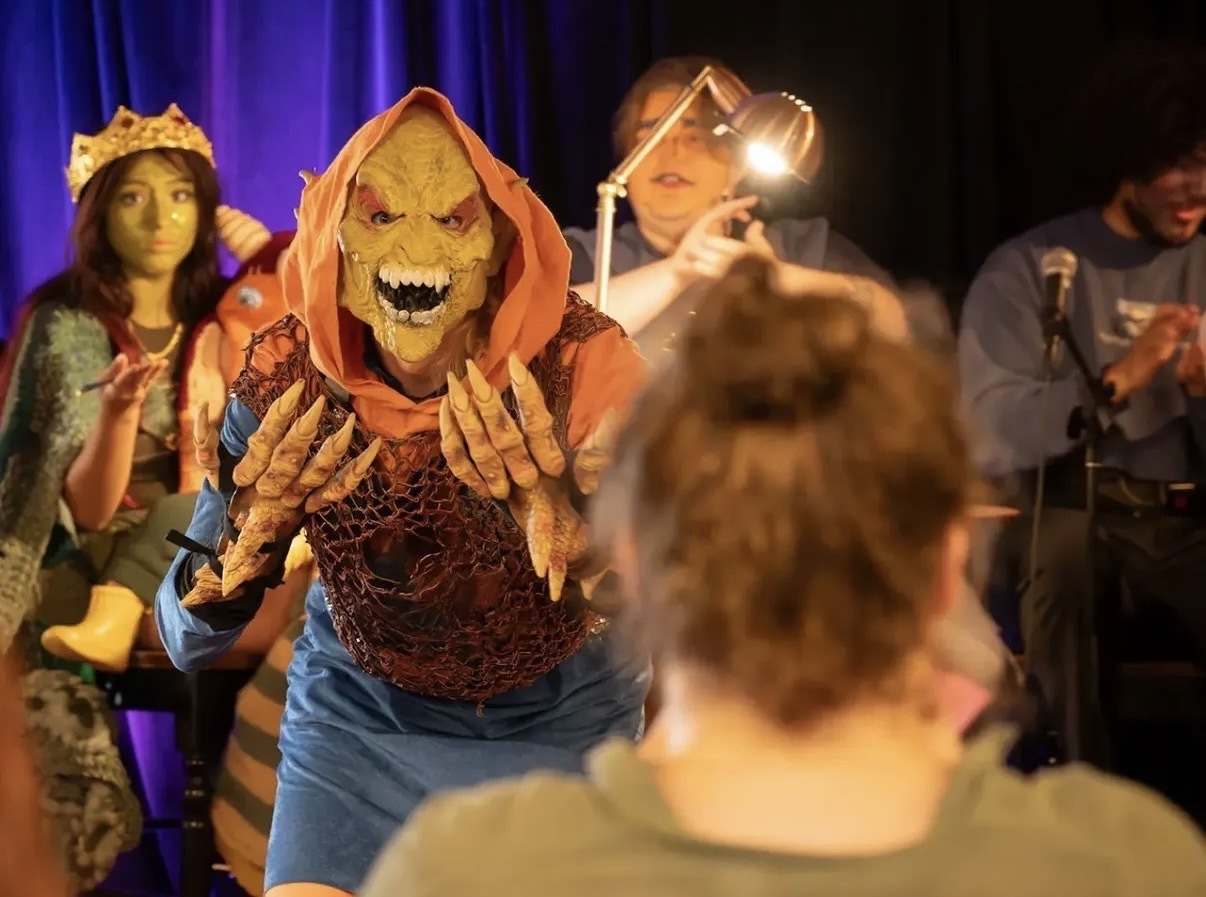 A person dressed as a goblin with other costumed people in the background. The back of an audience member's head is in the foreground.