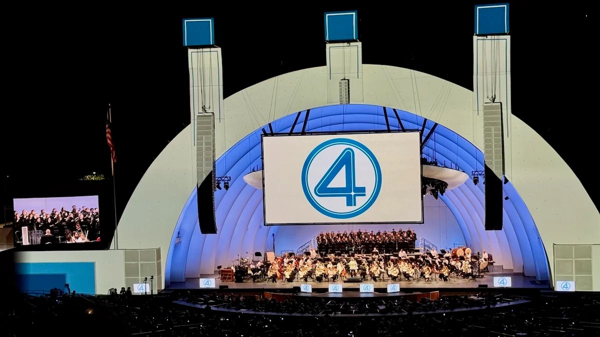 The big stage at the Hollywood Bowl. The arch is illuminated in white with a projection screen featuring the Fantastic 4 logo, a blue 4 in a circle. There is an orchestra and choir on stage, and they perform in front of a packed audience in the evening.