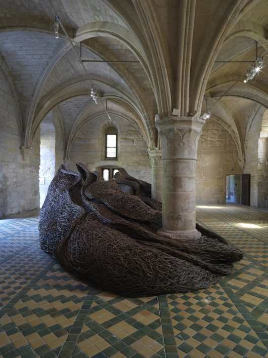 an organic sculptural form made from willow and tree branches wraps around a column in the room of an abbey.