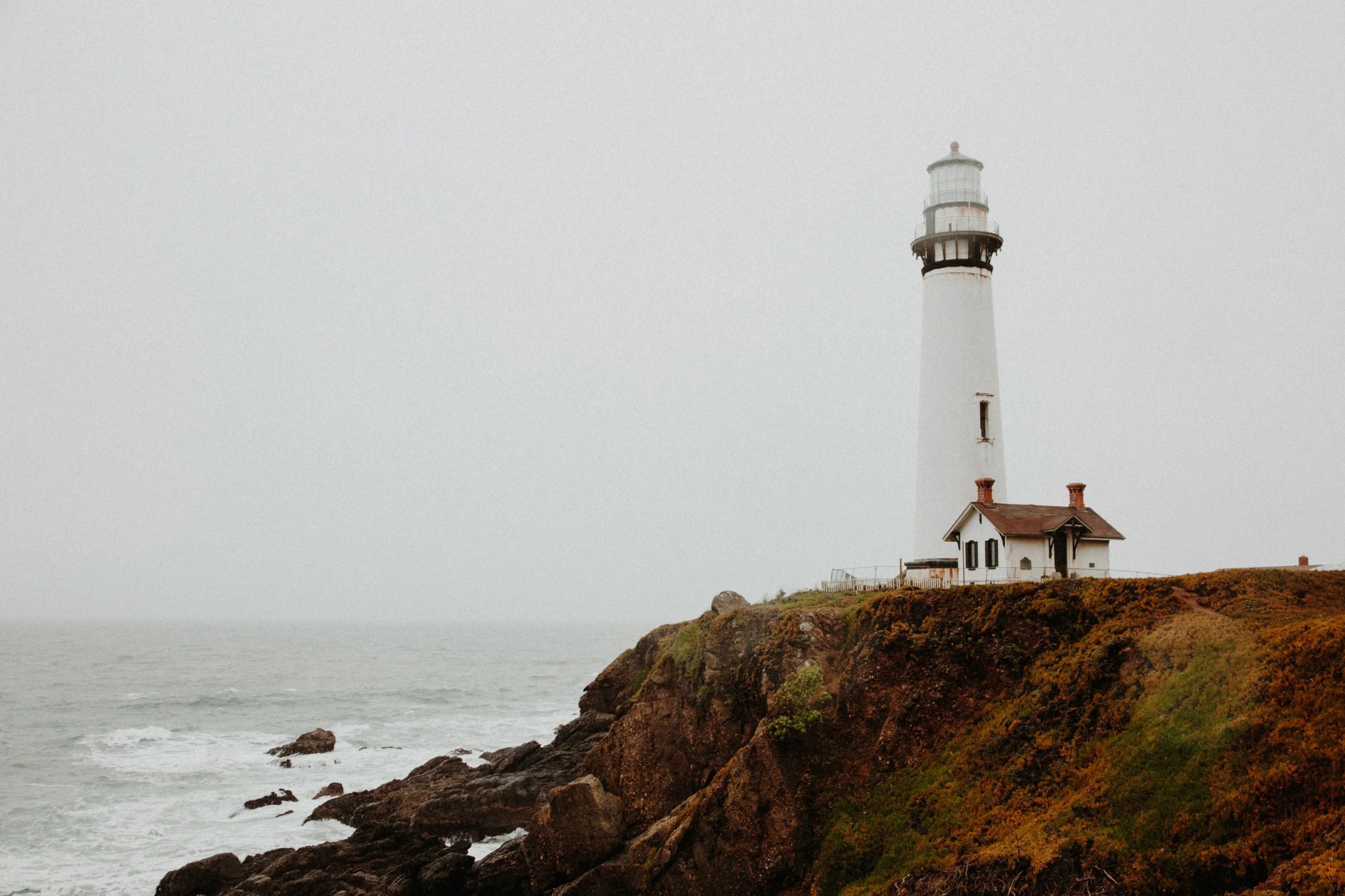 A white lighthouse stands on a grassy cliff above crashing ocean waves, with an overcast sky.