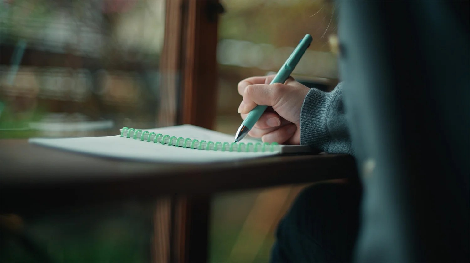 A person writing in a spiral notebook with a green pen near a large window.