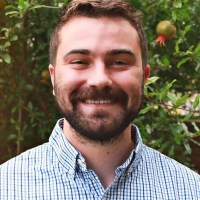 A bearded white man with chestnut colored trimmed hair and beard stands outside in a navy, light-blue, and white plaid button down shirt with only the top button remaining undone. Some teeth come through the beard to form a smile in front of a background of greenery. His right and left ears are slightly sunburned from the Texas summer. There is a ripe pomegranate growing from one of the trees in the background. The photo has been slightly altered to smooth out ridges on Nick's forehead and adjust the level of lighting.