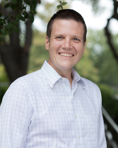 A white man in his early-40's, smiling with a blurred scene of trees in the background. He has blue eyes and short brown hair.