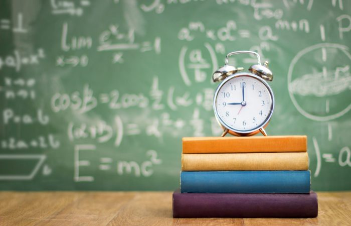 An alarm clock on top of books in front of a blackboard covered in white text
