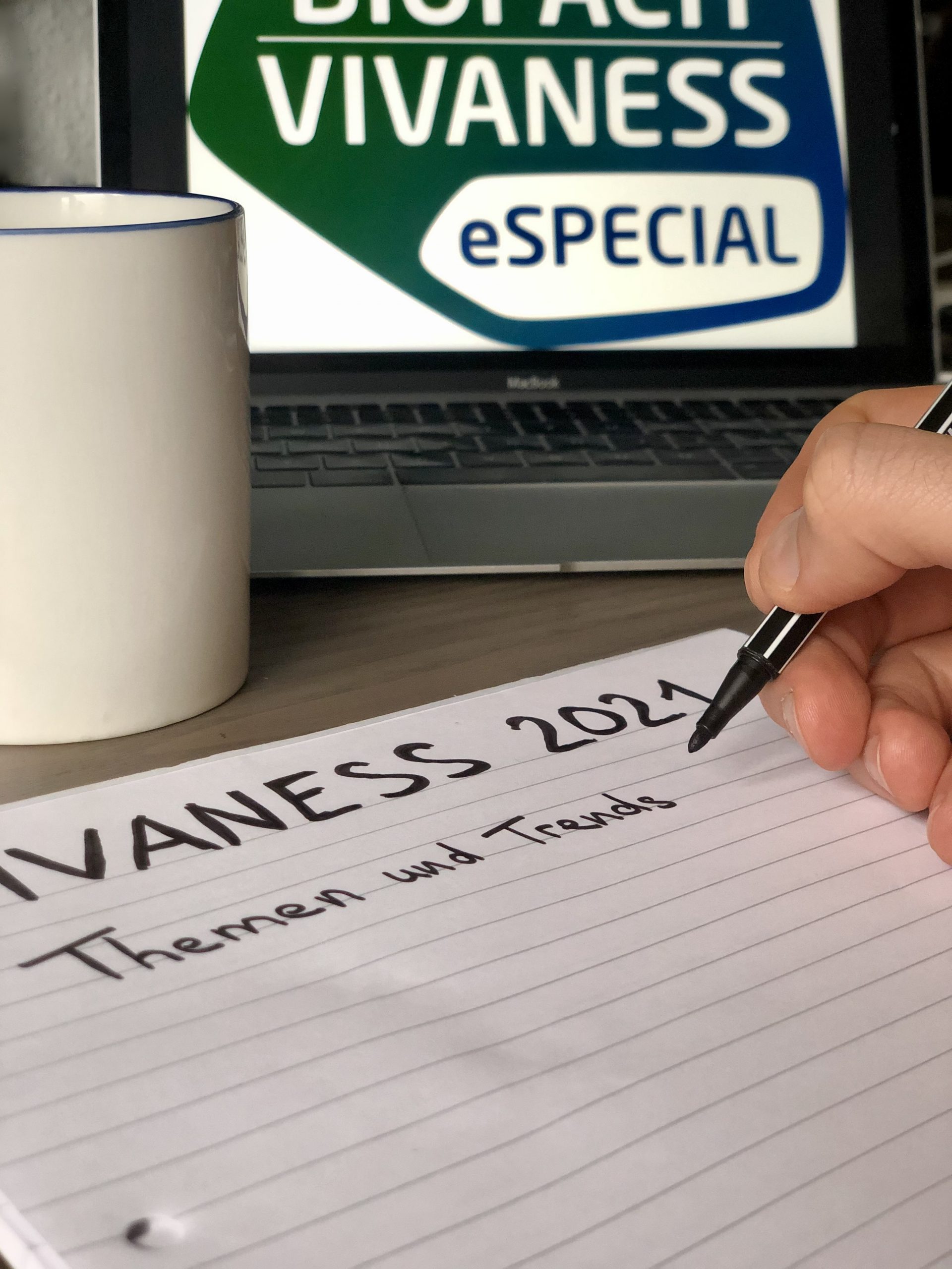 Auf dem Schreibtisch stehen eine weiße Tasse, ein Notizblock und Laptop mit Schriftzug VIVANESS 2021 eSPECIAL