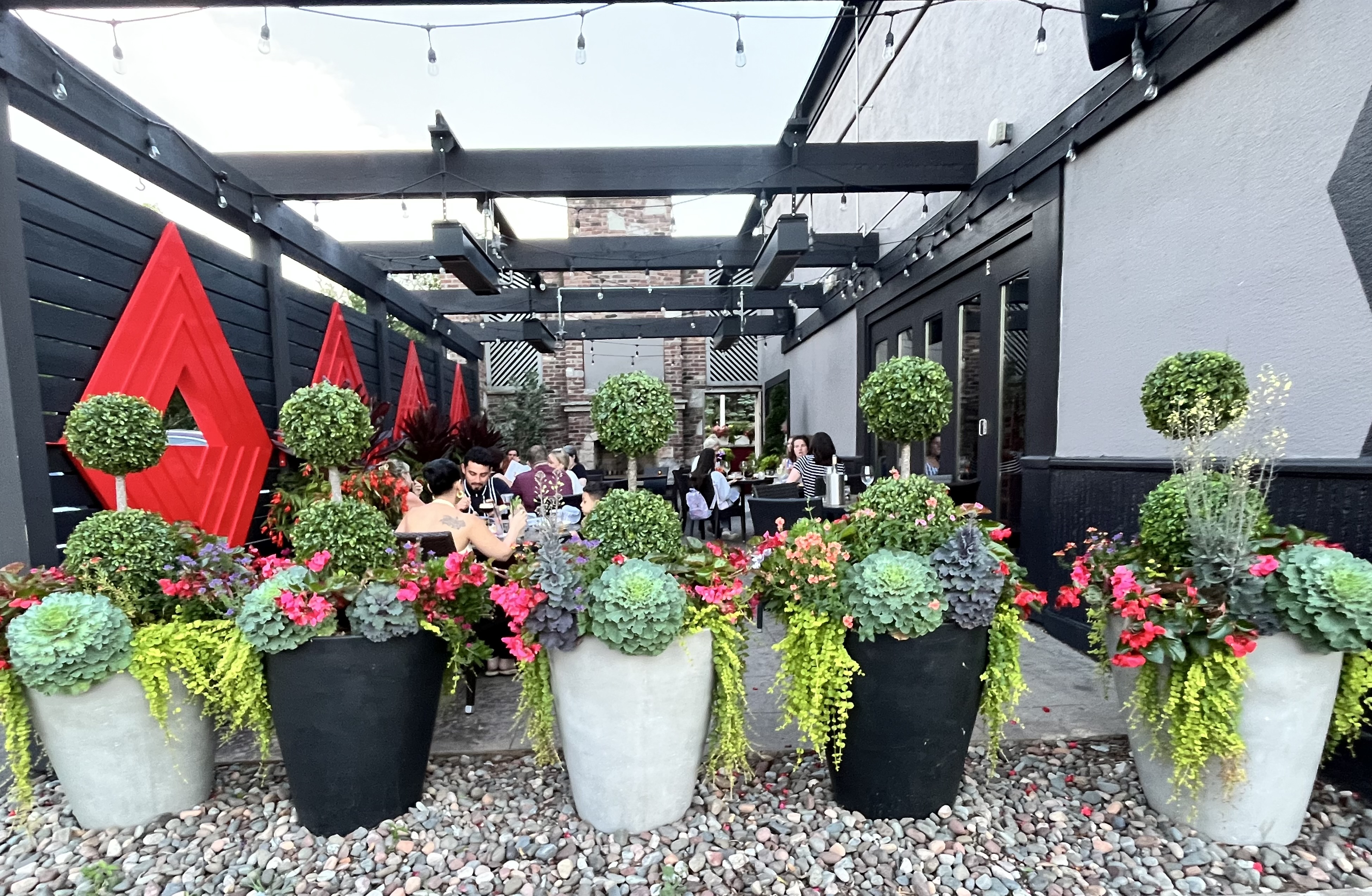 Patio planters at the edge of a patio that has edison lights and a black and red pergola