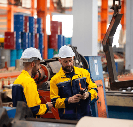 Two factory workers wearing yellow and navy uniforms and white helmets stand in an industrial setting, examining equipment and discussing something while surrounded by machinery and colorful storage bins.