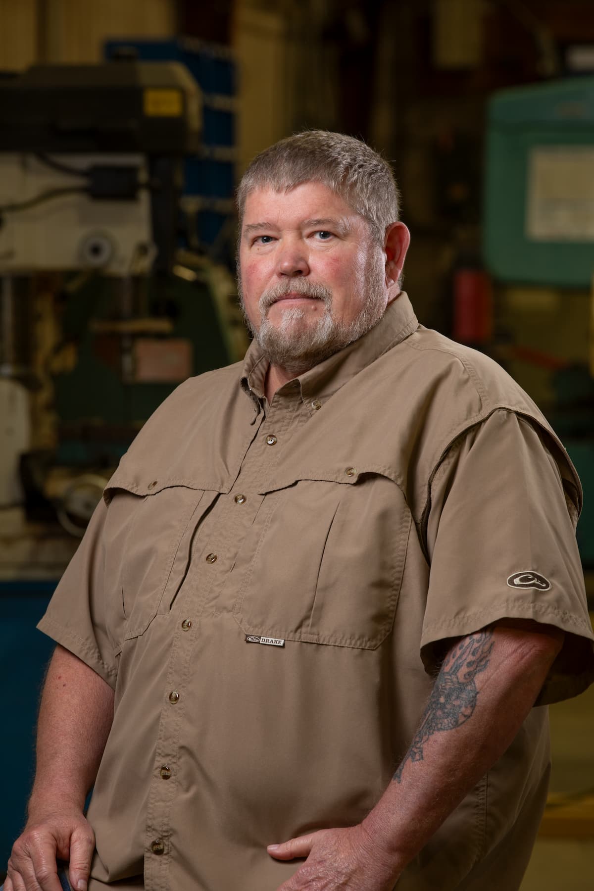 A man with short gray hair and a beard, wearing a tan button-up shirt, stands in an industrial setting. He has a tattoo on his right forearm and looks directly at the camera with a neutral expression.