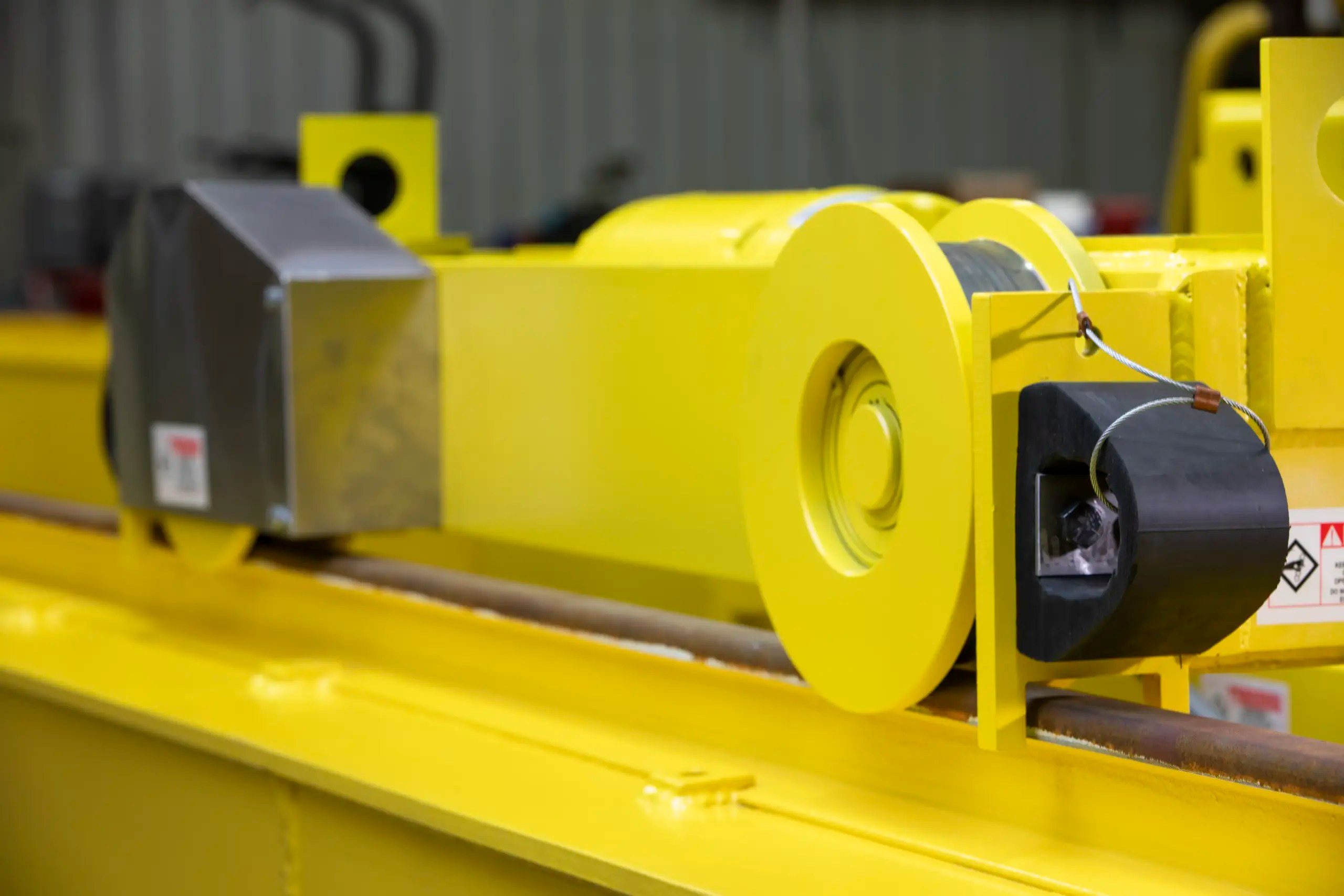 Close-up of a bright yellow industrial overhead crane mechanism with a metal cable wound around a spool and various mechanical components in a warehouse setting.