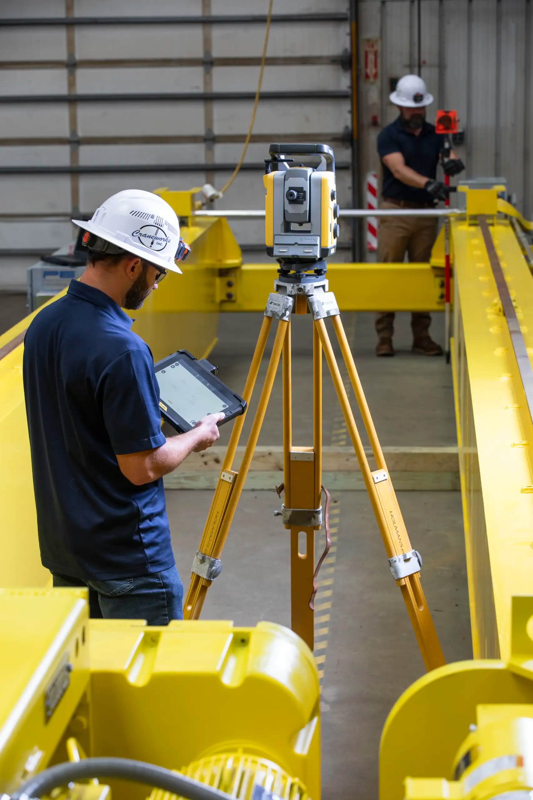Two workers in hard hats use surveying equipment and a tablet inside an industrial facility with yellow machinery and metal structures. One operates a tripod-mounted device while the other stands in the background.