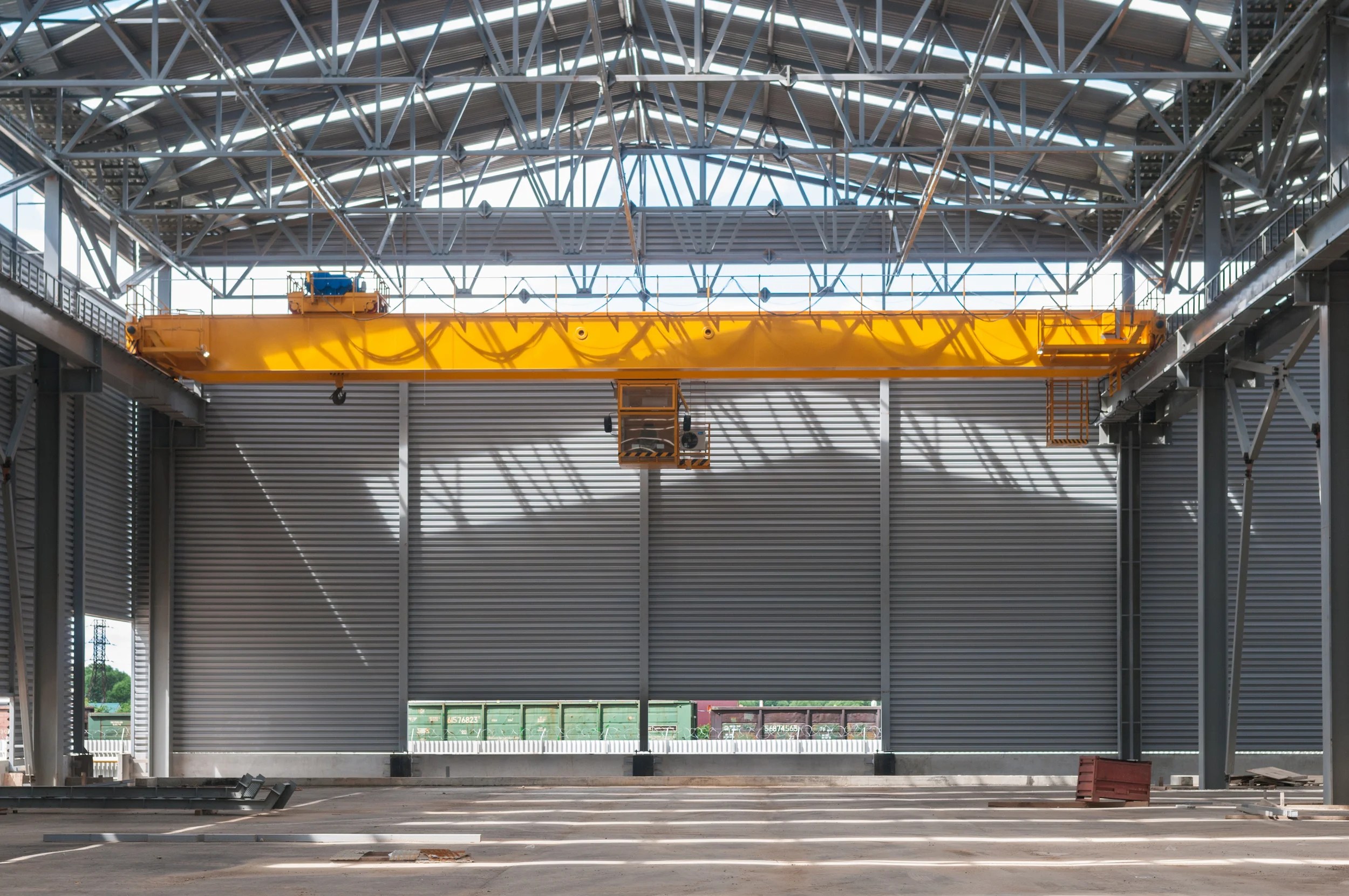 Interior of a large industrial warehouse with a high metal roof and steel beams, featuring a bright yellow overhead crane. The space is empty, with a few items on the concrete floor and sunlight streaming in.