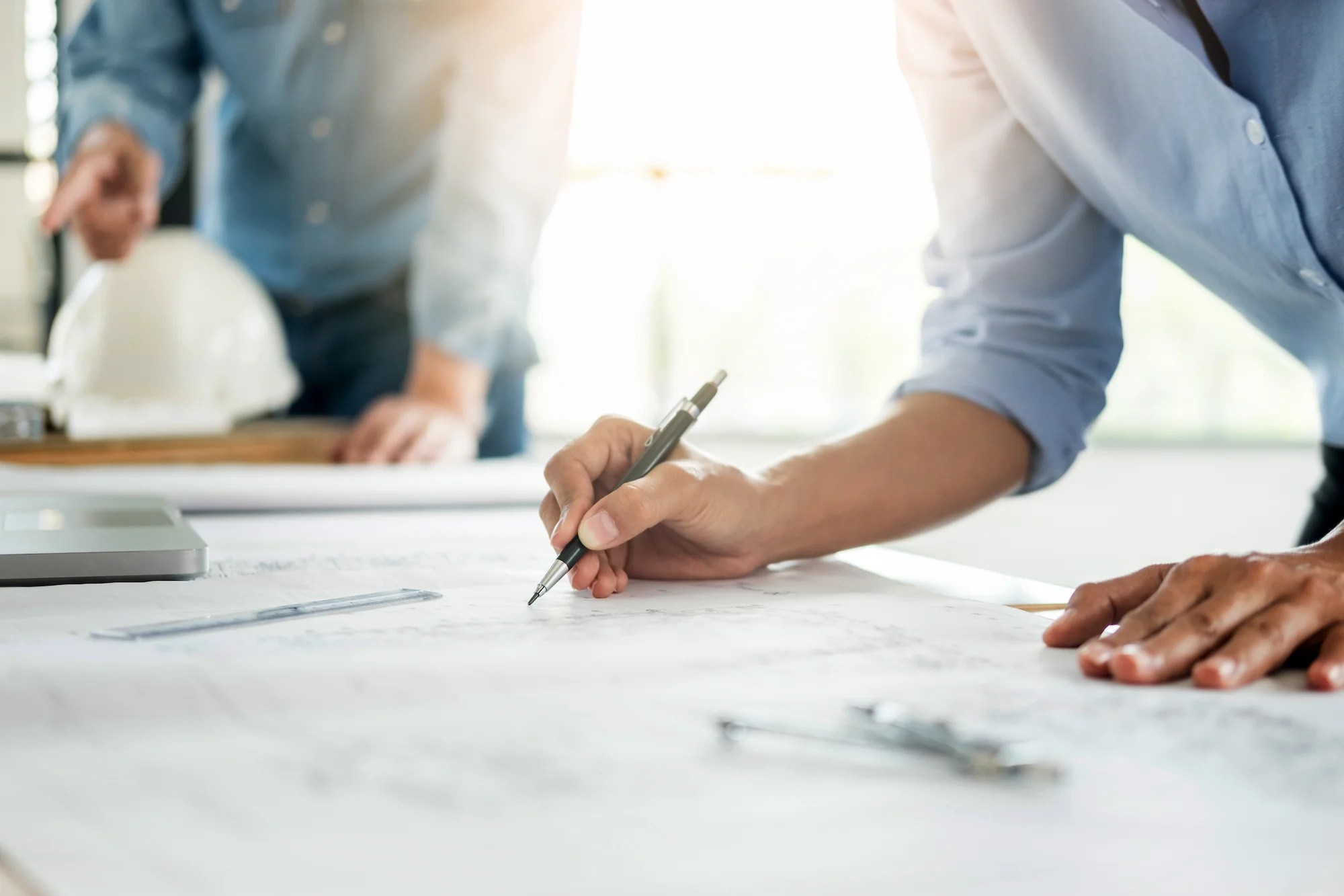 Close-up of a person’s hand drawing or writing on architectural blueprints on a desk, with another person and a white safety helmet blurred in the background. The scene is well-lit with natural light.